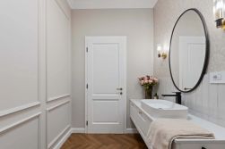 Wizualizacja jasnej łazienki w stylu klasycznym. Ściany zdobi sztukateria oraz tapeta. Na białej szafce stoi nablatowa umywalka z czarną baterią, a nad nią wisi owalne lustro w ciemnej ramie. Na podłodze ułożono parkiet w jodełkę.