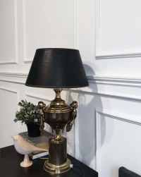 Klasyczne wnętrze z białą ścianą ozdobioną elegancką sztukaterią. Na ciemnym blacie stoi dekoracyjna lampa z brązowo-złotą podstawą w kształcie pucharu i czarnym kloszem. Obok widać małą roślinę w doniczce, książki oraz drewnianą figurkę ptaka.
