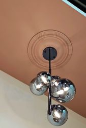 Nowoczesna lampa wisząca z kulistymi, szklanymi kloszami w kolorze chromu, zamontowana na czarnym stelażu. Lampa wisi na tle pudrowor&oacute;żowego sufitu, ozdobionego klasyczną, okrągłą rozetą sztukatorską. Widok od dołu.