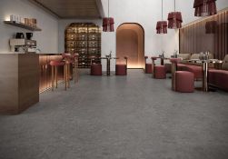 Wnętrze nowoczesnej restauracji z ciemnoszarą, nakrapianą podłogą. Po lewej stronie drewniany bar z hokerami i ekspresem. Po prawej stoliki z bordowymi pufami i kanapami na tle drewnianych lamel. W tle wysokie łuki, regał na wino i wiszące lampy.