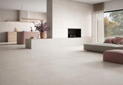 Jasny, przestronny salon z anneksem kuchennym i beżowymi płytami. Po prawej niska sofa, brudnor&oacute;żowy puf oraz wbudowany w ścianę kominek. W tle widać r&oacute;żową wyspę kuchenną, minimalistyczną lampę stojącą i wazon z kwiatami.