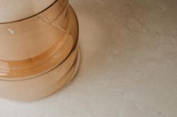 Fragment wazonu z miodowego, p&oacute;łprzezroczystego szkła o schodkowym kształcie, stojący w lewym rogu kadru. Naczynie umieszczono na jasnobeżowym, matowym blacie o delikatnej, nakrapianej strukturze kamienia.