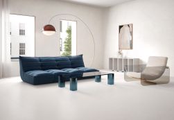 Nowoczesny salon z jasnymi płytami na podłodze i ścianie. W centrum stoi niska, pikowana sofa w kolorze granatowym oraz szklany stolik kawowy. Po prawej znajduje się designerski fotel, a nad sofą łukowa lampa stojąca z ciemnym kloszem.