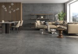 Nowoczesny salon z jadalnią, szarymi płytkami podłogowymi i ściennymi imitującymi beton, beżową sofą narożną, drewnianą ścianką działową, szklanym stolikiem kawowym i dużym oknem z widokiem na g&oacute;ry.