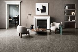 Elegancki, minimalistyczny salon z podłogą i obramowaniem kominka wykonanymi z ciemnego gresu lastryko z kolekcji Coem Porfirica Aglo Graphite. Centralny punkt stanowią dwa jasne fotele ustawione przy okrągłym drewnianym stoliku, a całość dopełnia nowocze