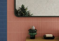 Zbliżenie na fragment ściany z wąskimi, pionowymi kafelkami typu lamelki w kolorze łososiowego r&oacute;żu i granatu. Na drewnianym blacie stoi szklany, zielony organizer ze szczoteczką oraz czarna mydelniczka. W lustrze widać odbicie rośliny.