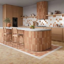 Nowoczesna kuchnia z wyspą kuchenną wyłożoną pionowymi terakotowymi płytkami na podłodze mniejsze kwadratowe kafelki w beżowym i terakotowym kolorze ze ścianą nad blatem w białe i terakotowe kafelki wiklinowe hokery i wbudowane czarne sprzęty AGD.