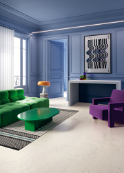 Salon, ściany w kolorze niebieskim, na środku zielona kanapa i zielony stolik, na podłodze dywan, obok fioletowy fotel, na podłodze płytki Superclassica SCW Honed 60x120 płytki imitujące kamień