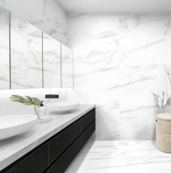 Łazienka, na białym blacie dwie umywalki, pod spodem czarna półka, duże lustro na ścianie, w rogu kosz na pranie, ściana i podłoga wyłożone płytkami Nuba Blanco 50x100 płytka imitująca marmur