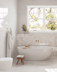 Łazienka, przy ścianie wanna wolnostojąca, duże okno, przy wannie małe drewniane krzesło z ręcznikiem, na wieszakach ściennych białe ręczniki kąpielowe, podłoga wyłożona białymi matowymi płytkami, ściana przy wannie wyłożona dekoracyjnymi płytkami z kolek