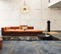 Salon, na środku duża pomarańczowa kanapa, na podłodze przy kanapie dywan, mały okrągły czarny puf, na podłodze niebieskie płytki, ściana wyłożona Flamed Ice Natural Rect. 99,55x99,55 płytki imitujące metal