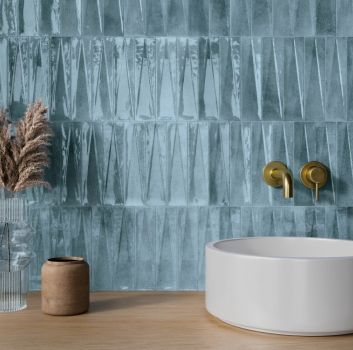 Ściana w łazience wyłożona niebieskimi cegiełkami z drewnianym blatem i białą, okrągłą umywalką, złotą baterią podtynkową i ozdobami