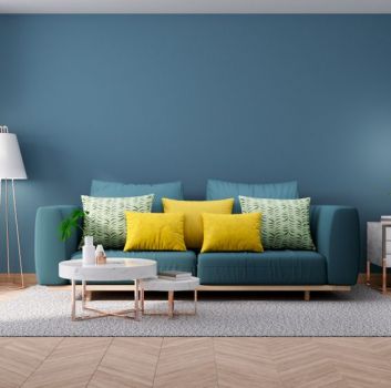 Salon z drewnopodobną podłogą w jodełkę i niebieską ścianą, niebieską kanapą z kolorowymi poduszkami, fotelem, lampą stojącą i kwiatem w donicy