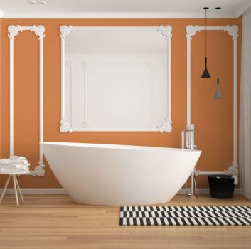 Łazienka z drewnianą podłogą i pomarańczowymi ścianami, białą wanną wolnostojącą i czarno-białym dywanikiem, ścienną umywalką i lustrem w białej, ozdobnej ramie
