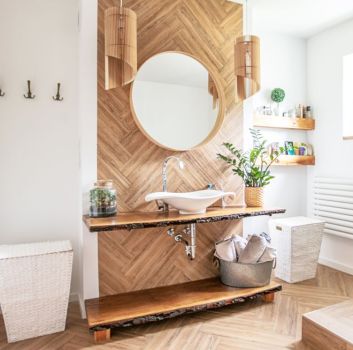 Przytulna łazienka wyłożona płytkami drewnopodobnymi z umywalką nablatową, okrągłym lustrem, dwoma lampami wiszącymi, zabudowaną wanną i koszem wiklinowym