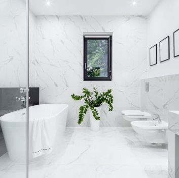 Jasna łazienka wyłożona białymi płytkami imitującymi marmur z szarymi żyłami z białą wanną owalną, wiszącą miską WC i bidetem, kabiną prysznicową, oknem i kwiatem w donicy i trzema obrazkami na ścianie
