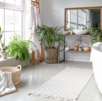 Biała łazienka z dużą wanną, dywanikiem na podłodze, szafką z dwoma półkami a akcesoriami i umywalką nablatową, dużym lustrem, koszem wiklinowym, wysoką drabiną z szlafrokiem, dużym oknem i dużą ilością kwiatów