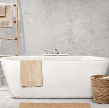 Fragment minimalistycznej łazienki wyłożonej szarymi płytkami imitującymi beton z białą wanną wolnostojącą z przewieszonym beżowym ręcznikiem, brązowym dywanikiem, koszem wiklinowym i drabiną z ręcznikami
