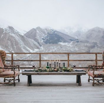 Drewniany taras z zastawionym stołem i dwoma fotelami w stylu vintage z rozległym widokiem na góry zimą