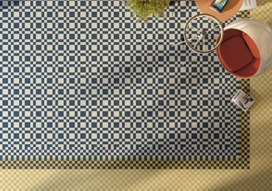 Wizualizacja przedstawia widok z g&oacute;ry na geometryczną podłogę z tych płytek, uzupełnioną o designerski, nowoczesny fotel z rudym środkiem, szklany stolik oraz leżący na ziemi album o architekturze.