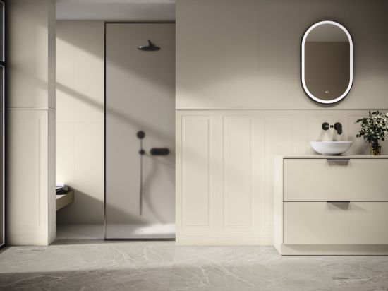 Jasna, nowoczesna łazienka w odcieniach beżu. Beżowa szafka z białą umywalką, owalne, podświetlane lustro i czarna bateria ścienna. Ściany ze sztukaterią, w tle kabina prysznicowa walk-in.