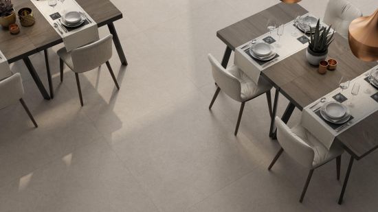 Widok z g&oacute;ry na jasne, matowe płyty podłogowe w jadalni lub restauracji. Po obu stronach kadru stoją ciemne drewniane stoły z kompletną zastawą i bieżnikami, przy kt&oacute;rych ustawiono eleganckie, tapicerowane krzesła w odcieniu złamanej bieli.