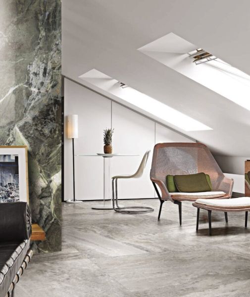 Jasne poddasze ze ścianą z zielonego marmuru Florim Heritage Luxe Emerald i szarymi płytkami Cloud na podłodze. Wnętrze zdobią designerskie meble: pleciony fotel, biały stolik i lampa stojąca.