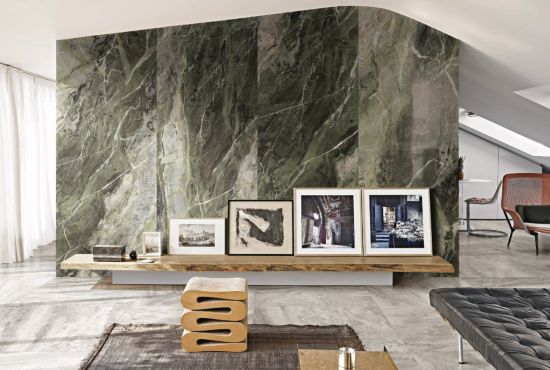 Wizualizacja salonu z dekoracyjną ścianą z zielonego marmuru Florim Heritage Luxe Emerald. Na drewnianej p&oacute;łce stoją oprawione grafiki, a wnętrze dopełnia niska pufa i szara, kamienna podłoga.