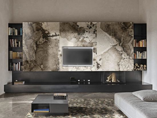 Nowoczesny salon z telewizorem na tle ściany z płyt Florim Heritage Luxe Flame o wzorze brązowo-beżowego marmuru. Widoczna czarna niska zabudowa, regały na książki, kominek i szara sofa.