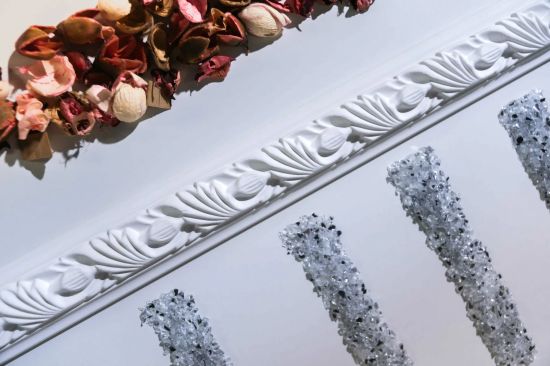 Zbliżenie na dekoracyjny, biały gzyms z klasycznym motywem roślinnym. Nad nim widoczna jest kompozycja z suszonych płatk&oacute;w kwiat&oacute;w w odcieniach r&oacute;żu i brązu, a poniżej znajdują się pionowe pasy zdobione błyszczącym, drobnym kryształem.