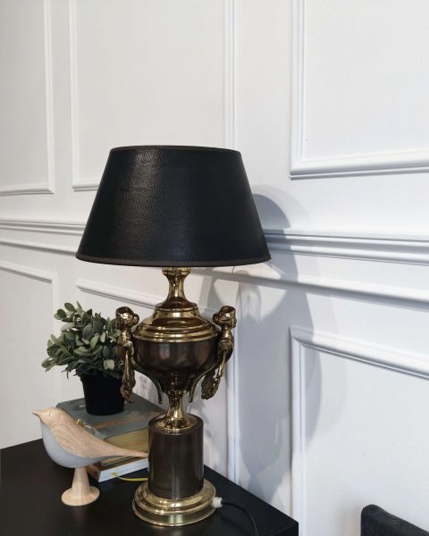 Klasyczne wnętrze z białą ścianą ozdobioną elegancką sztukaterią. Na ciemnym blacie stoi dekoracyjna lampa z brązowo-złotą podstawą w kształcie pucharu i czarnym kloszem. Obok widać małą roślinę w doniczce, książki oraz drewnianą figurkę ptaka.