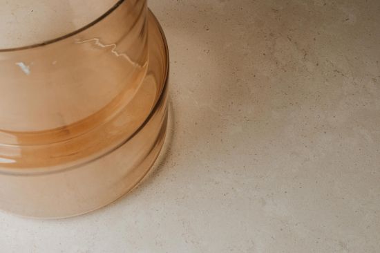 Fragment wazonu z miodowego, p&oacute;łprzezroczystego szkła o schodkowym kształcie, stojący w lewym rogu kadru. Naczynie umieszczono na jasnobeżowym, matowym blacie o delikatnej, nakrapianej strukturze kamienia.