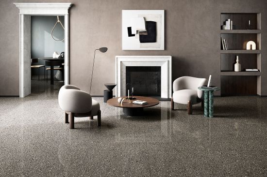 Elegancki, minimalistyczny salon z podłogą i obramowaniem kominka wykonanymi z ciemnego gresu lastryko z kolekcji Coem Porfirica Aglo Graphite. Centralny punkt stanowią dwa jasne fotele ustawione przy okrągłym drewnianym stoliku, a całość dopełnia nowocze