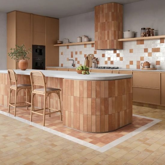Nowoczesna kuchnia z wyspą kuchenną wyłożoną pionowymi terakotowymi płytkami na podłodze mniejsze kwadratowe kafelki w beżowym i terakotowym kolorze ze ścianą nad blatem w białe i terakotowe kafelki wiklinowe hokery i wbudowane czarne sprzęty AGD.