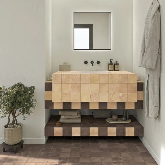 Nowoczesna łazienka z podwieszanymi meblami wyłożonymi beżowymi i ciemnobrązowymi kwadratowymi płytkami czarnym lustrem nad umywalką rośliną doniczkową po lewej i szlafrokiem po prawej stronie.